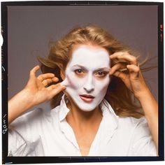 a woman with white face paint holding her hands up to her head and looking at the camera