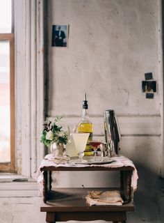 a table with bottles and glasses on it in front of a window next to an open book