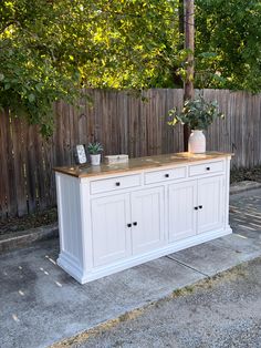 a white buffet table with two potted plants on top in front of a wooden fence