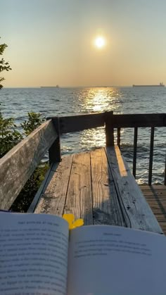 an open book sitting on top of a wooden bench next to the ocean at sunset