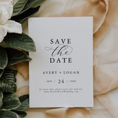 save the date card with white flowers and greenery next to it on a bed