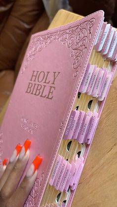 a woman's hand holding a pink bible with orange nails