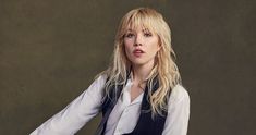 a woman with long blonde hair sitting on a chair in front of a wall wearing a white shirt and black vest