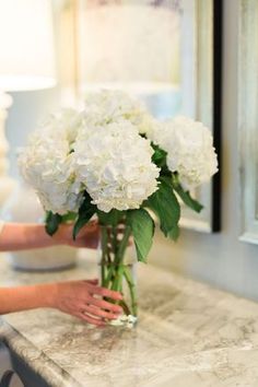 a woman arranging white flowers in a vase on a marble counter top with a mirror behind her