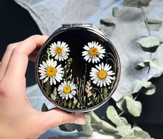 a hand holding a compact mirror with daisies on it and greenery in the background