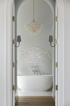 a white bath tub sitting under a chandelier next to a doorway with two lamps