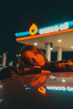 a woman leaning on the hood of a car in front of a gas station at night