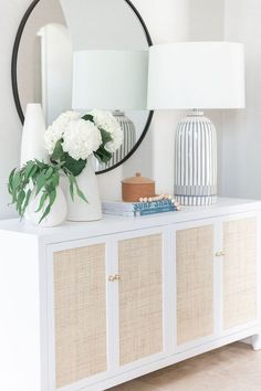 a white sideboard with flowers on top and a mirror in the corner behind it