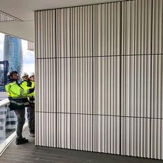 two men in yellow vests standing next to each other near a wall with vertical slats on it