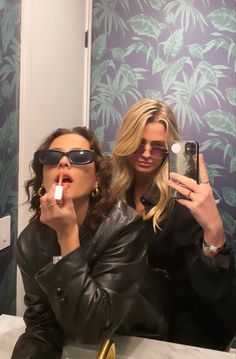 two women in sunglasses are brushing their teeth while taking a selfie with an iphone