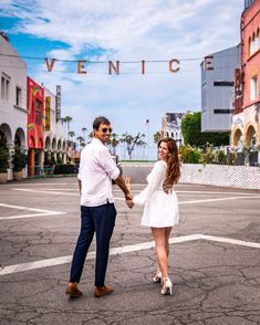 a man and woman holding hands in the middle of an empty parking lot with buildings behind them