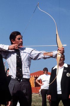 a man holding a bow and arrow in front of other men standing around with their backs to the camera