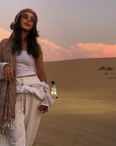 a woman standing in the desert wearing white pants and a scarf around her neck with two people behind her