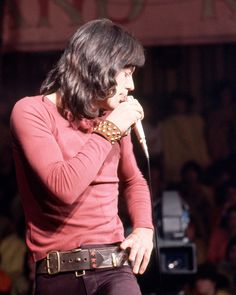 a man with long hair standing in front of a microphone and holding his hand to his mouth