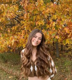 a woman standing in front of a tree with long brown hair and wearing a sweater