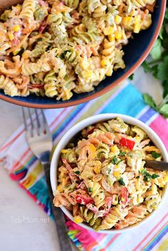 two bowls filled with pasta salad on top of a colorful table cloth next to a wooden spoon