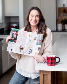 a woman standing in a kitchen holding up a magazine and a cup with coffee on it