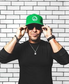 a man in black shirt and green hat holding his hands up to his face with white brick wall behind him