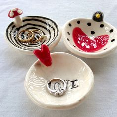 three ceramic bowls with rings in them on a white tableclothed surface, one has a ring holder and the other is a bowl that says love