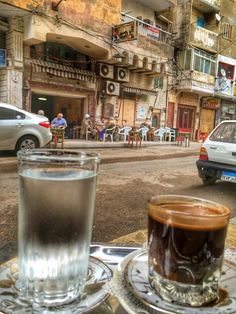 two cups of coffee sit on a table in front of an old building and parked cars