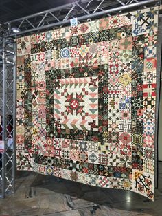 a large quilt hanging from the ceiling in front of a metal stand with several different designs on it