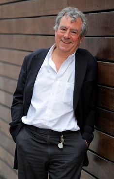 a man leaning against a wooden wall with his hands in his pockets and smiling at the camera