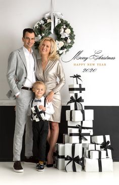 a man and woman standing next to a small boy in front of christmas presents with a wreath on the wall behind them