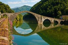 an old stone bridge spanning the width of a river with flowers growing on it and mountains in the background