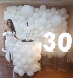 a woman standing next to a table with white balloons on it and a bottle of booze