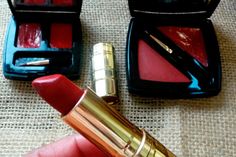 a person holding a red lipstick in their hand next to some other items on a table