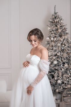 a pregnant woman in a white gown standing next to a christmas tree with her hands on her belly