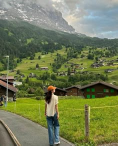 a woman standing on the side of a road next to a lush green hillside covered in snow