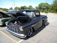 an old black truck parked in a parking lot next to another car with its hood open