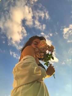 a woman in a white dress holding flowers up to her face and looking at the sky