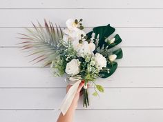 a person holding a bouquet of flowers in front of a white wall with palm leaves