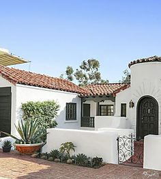 a white house with black doors and red tile on the front yard, surrounded by greenery