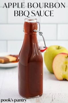 an apple cider next to two apples on a white table with a glass carafe in the foreground