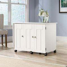 a white cabinet with two doors on wheels in front of a blue wall and wooden floor