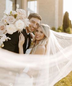 a bride and groom pose for a photo in front of their wedding venue with the veil blowing in the wind