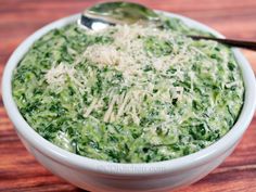 a white bowl filled with spinach and cheese on top of a wooden table next to a spoon