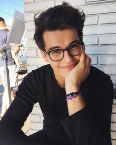 a man wearing glasses sitting at a table in front of a white brick wall with his hand on his chin