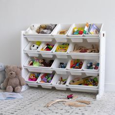 a toy storage unit filled with lots of toys next to a teddy bear on the floor