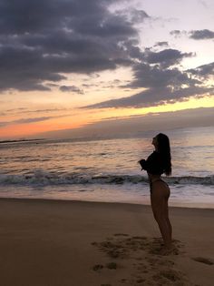 a woman standing on top of a sandy beach next to the ocean at sunset with clouds in the sky