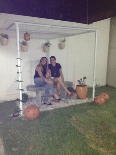 two women are sitting on a bench in the yard with potted plants and pumpkins