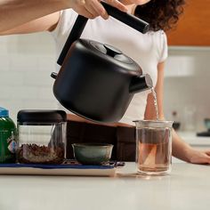 a woman pours water into a teapot while standing in the kitchen with other ingredients