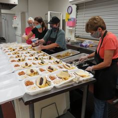 some people are preparing food at a buffet table with white trays on it and one person in an orange shirt is wearing a face mask