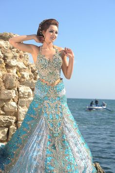 a woman in a blue and gold dress standing on rocks near the ocean with her hands behind her head