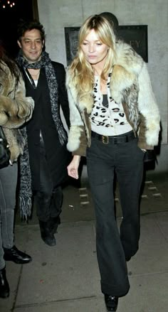 a woman walking down a sidewalk next to a man in a fur coat and black pants