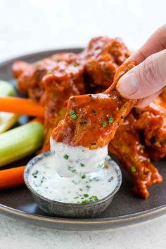 a hand dipping sauce on top of chicken wings with carrots and celery