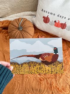 a person holding up a card with a pheasant on it and pumpkins in the background